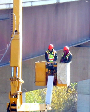 2 men in a bucket alongside a bridge