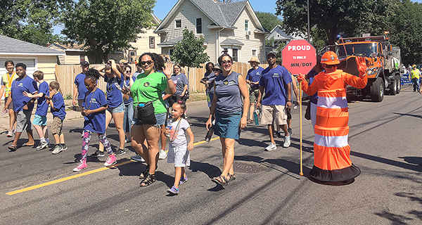 Group of MnDOT employees walking in front of orange snowplow as part of Rondo Days parade.