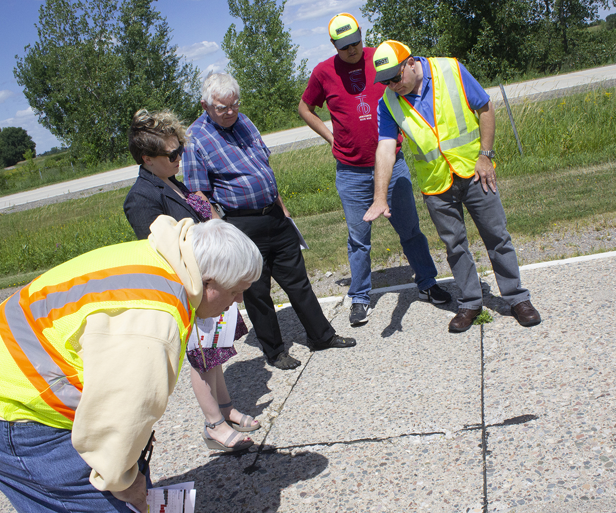 A man in a yellow safety vests gestures toward a section of concrete roadway, as others look at it too.