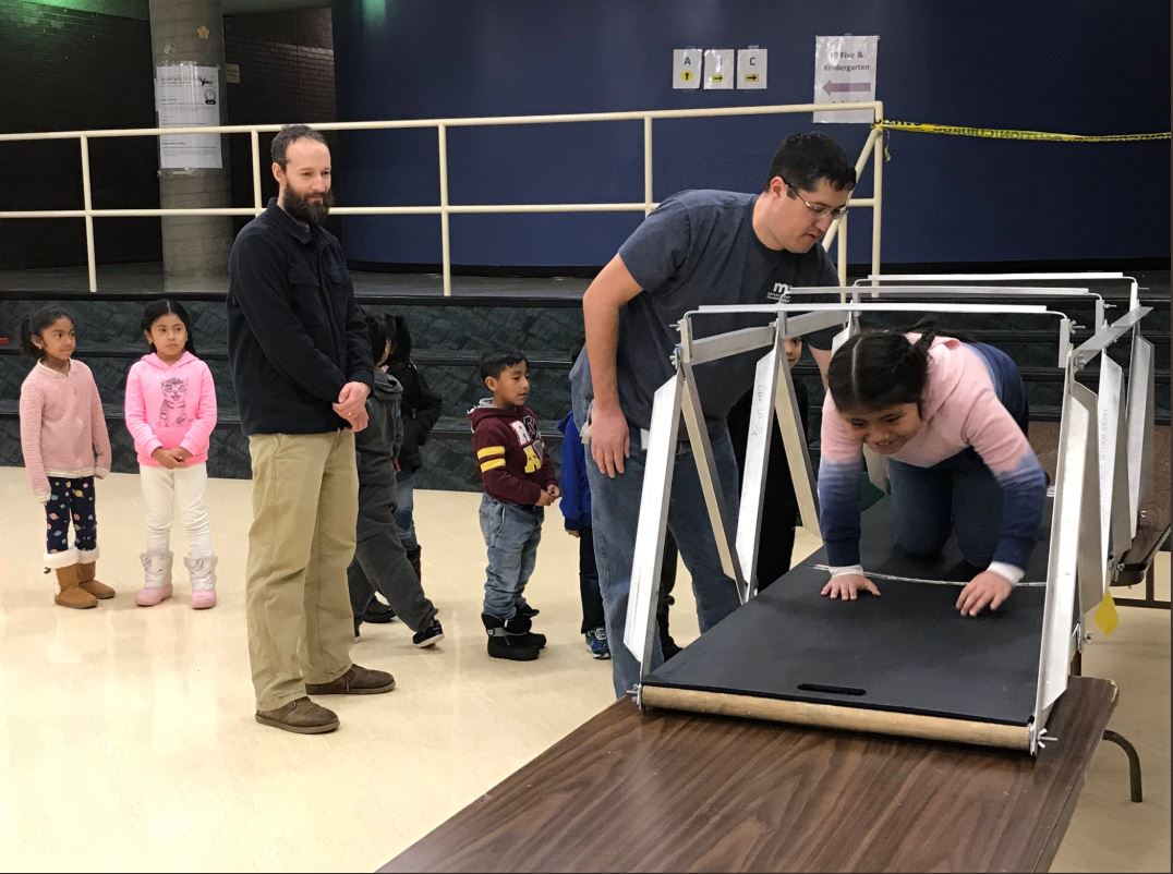 Photo: a MnDOT staff member helps a child crawl across a model bridge suspended between two tables.