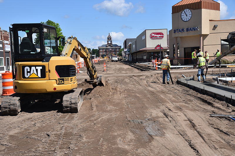 Two crew members watch an excavator dig on a torn up road site near buildings on highway 55