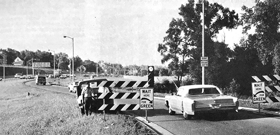 Black and white photograph showing an old car waiting to enter the freeway. The sign next to the car says wait here for green light