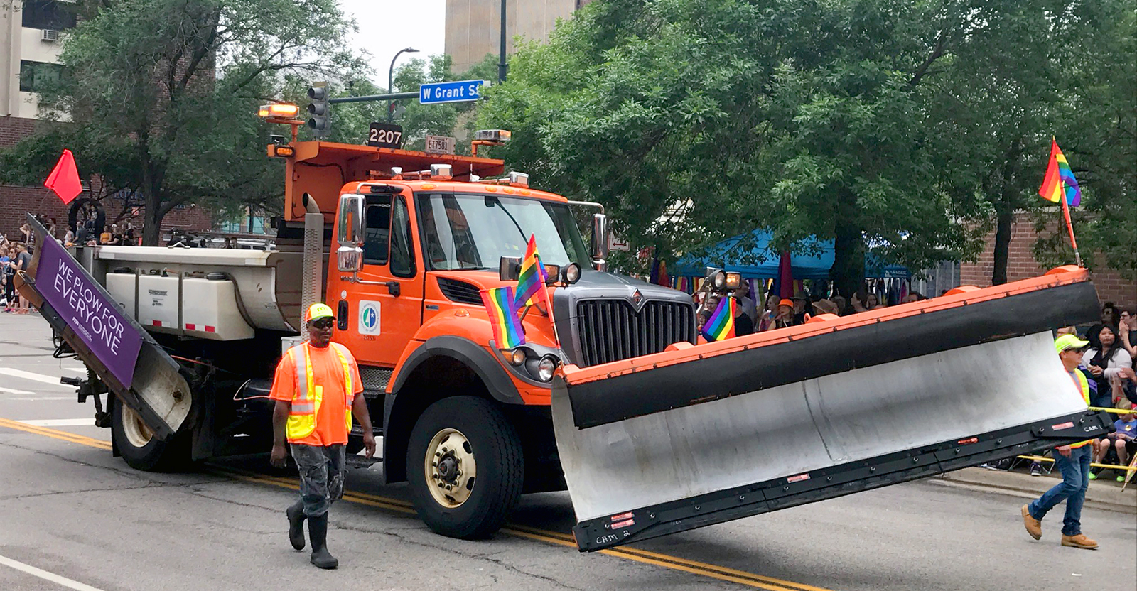 Picture of a plow truck driving through downtown Minneapolis. The side blade features a banner which reads we plow for everyone