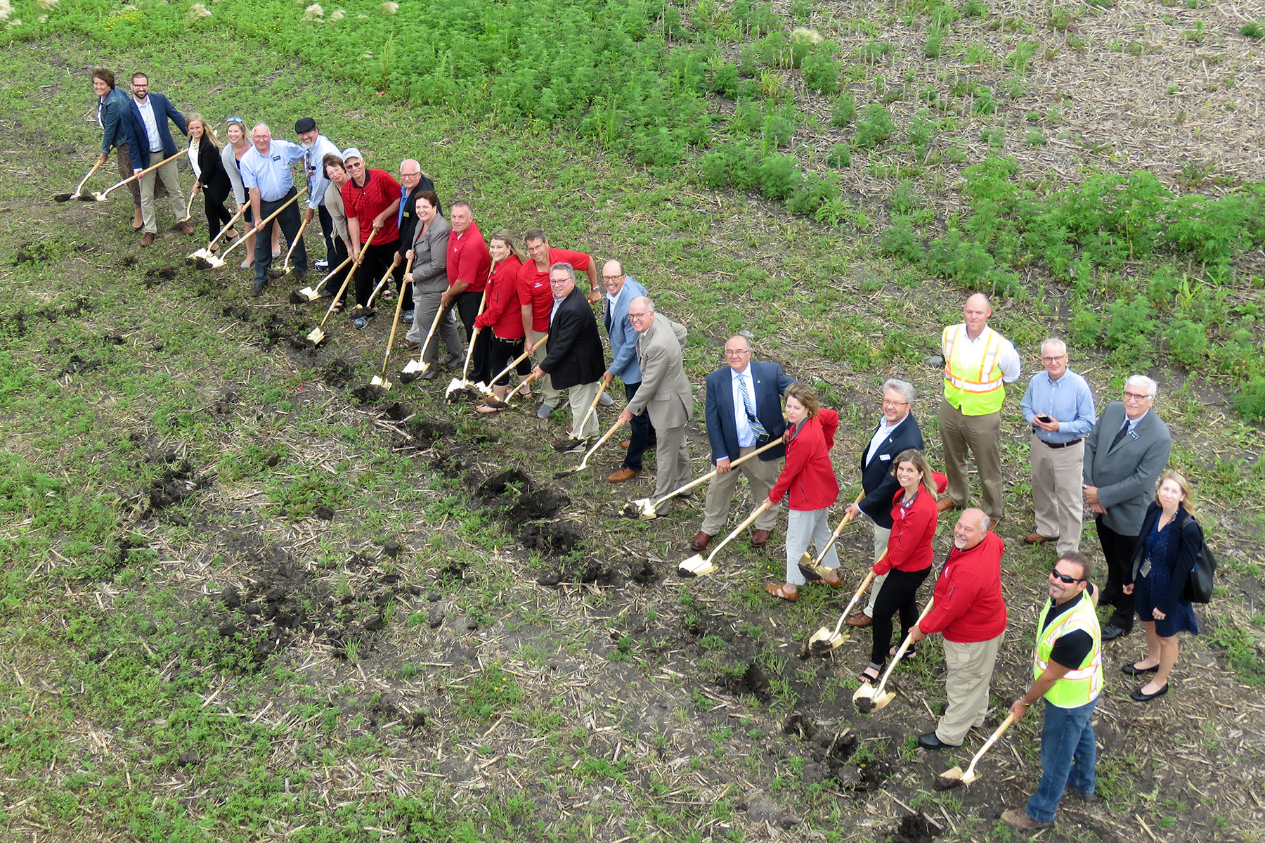 Nearly 20 people are pictured in a line, sticking gold painted shovels into the earth and looking up at the drone which is taking the picture above them.