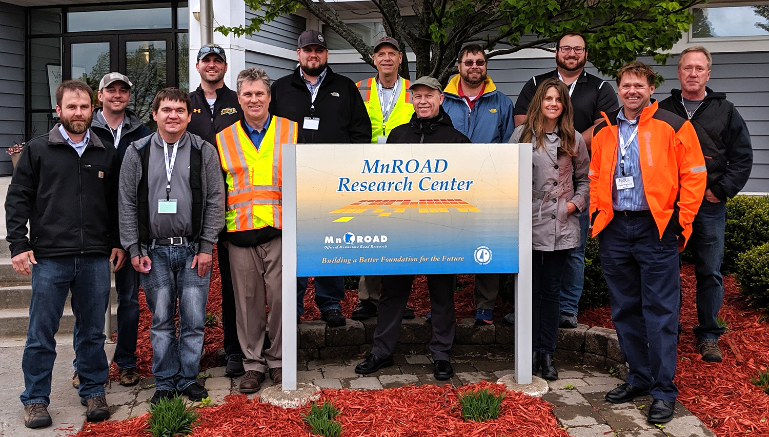 Group of people posing outdoors around a sign which reads MnROAD research center