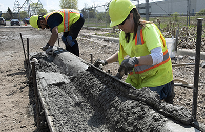 Two women working with concrete, forming it into the shape of a curb.