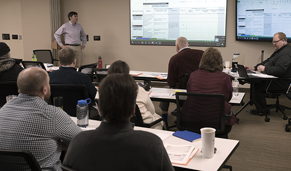 Seven seated people listen to two trainers. Two screens at front of room display info in a table format.