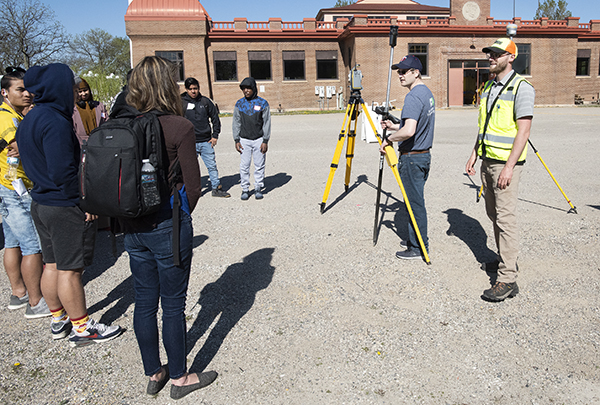 Two MnDOT employees talking to students in an outdoor setting. The employees are standing near surveying equipment.