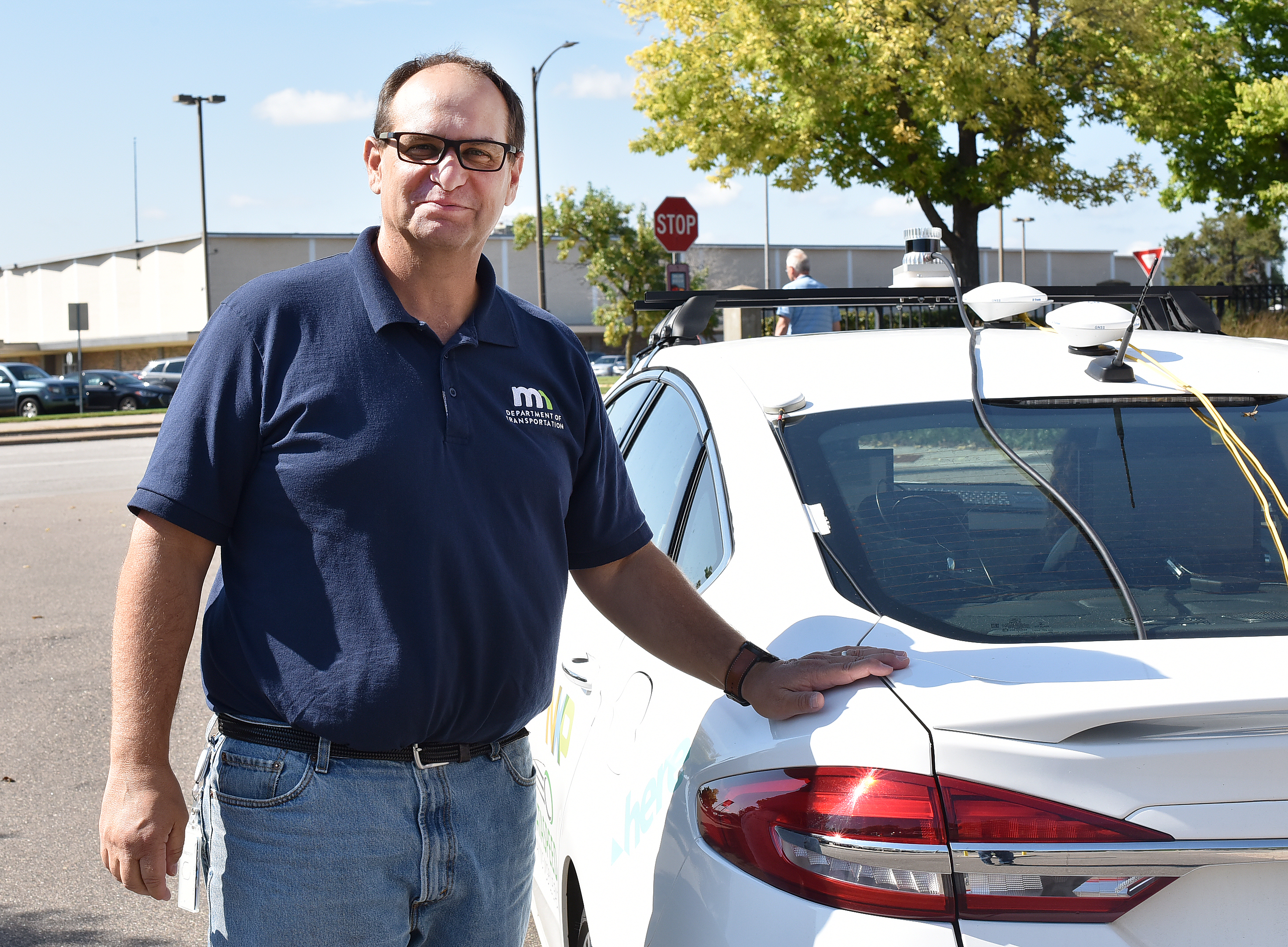 A man poses next to a car. The car has several external sensors mounted on the roof