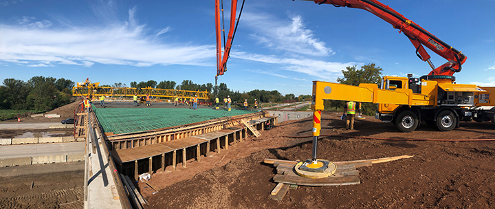 A large yellow truck extends a red boom arm over a partially finished bridge project as crews pour concrete.