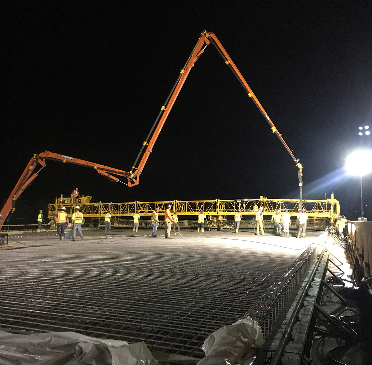 A construction crew works at night, while a large crane-type device looms over them, pouring concrete.