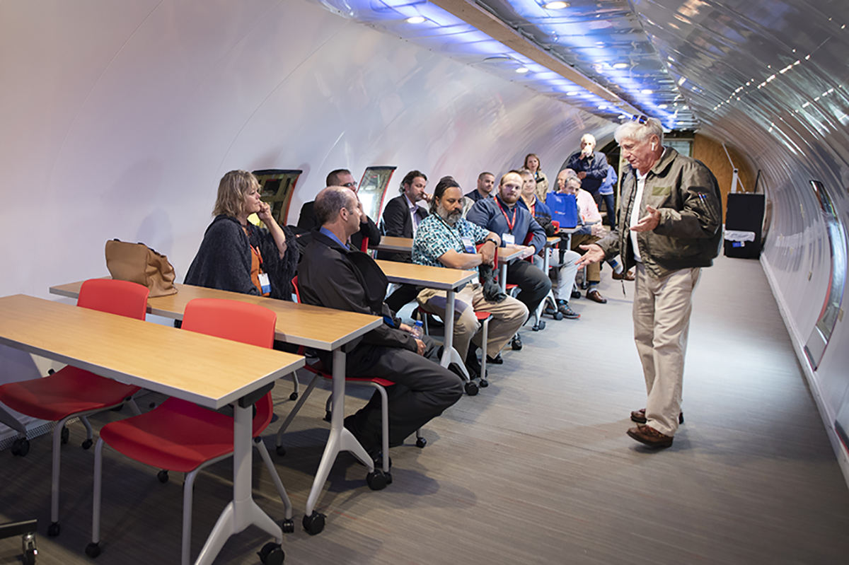 A standing man speaks to people seated at desks inside of a converted airplane fuselage.