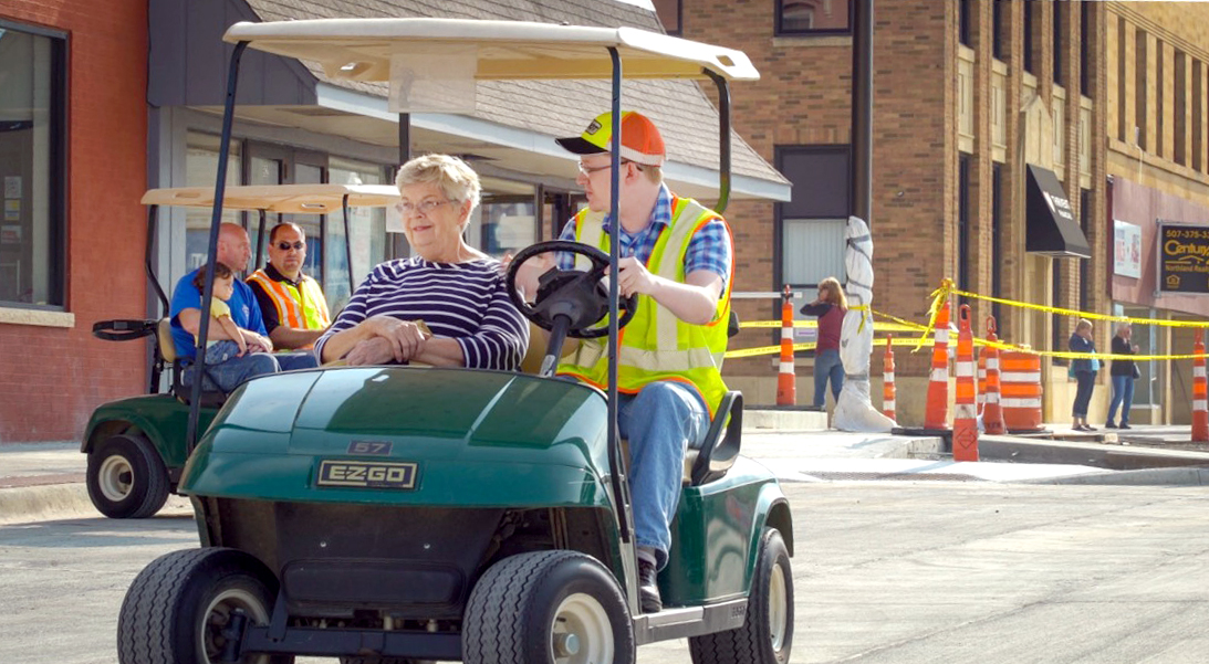 A man in a safety vest and hard hat drives a golf cart down a city street, while listening to his passenger.