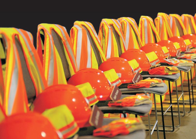 Photo: Orange helmets and vests draped on a row of chairs, each with the name of a fallen worker on them.