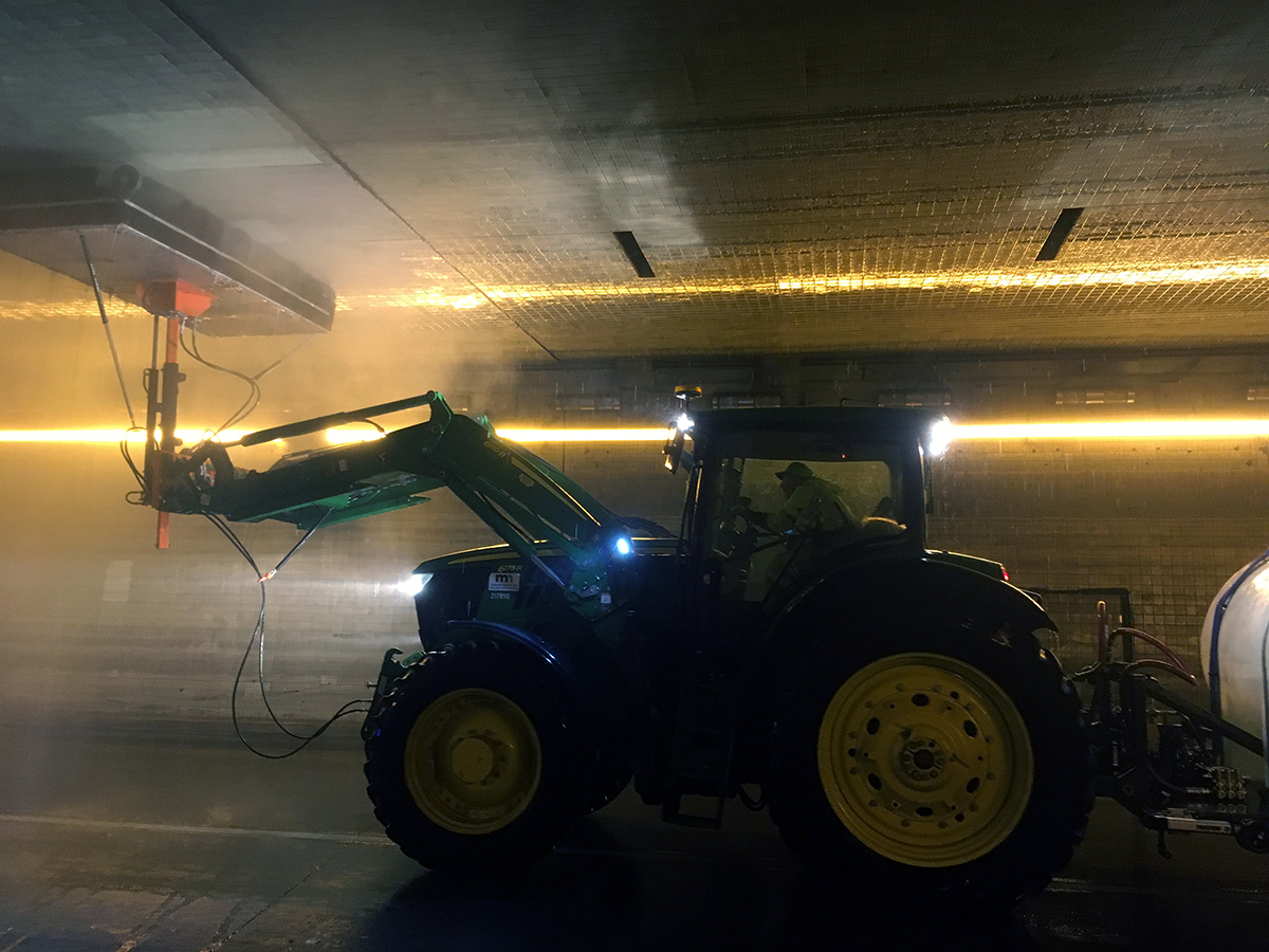 Photo: a John Deere tractor with a large spraying device mounted on the front arm, washing a tunnel ceiling