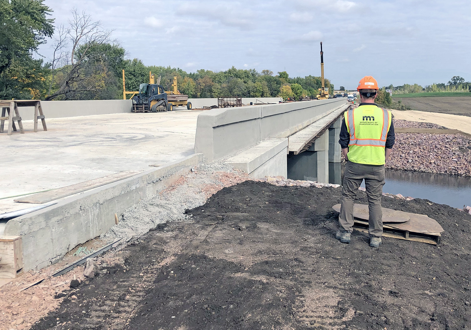 Photo: a worker in a MnDOT vest standing near a bridge under construction