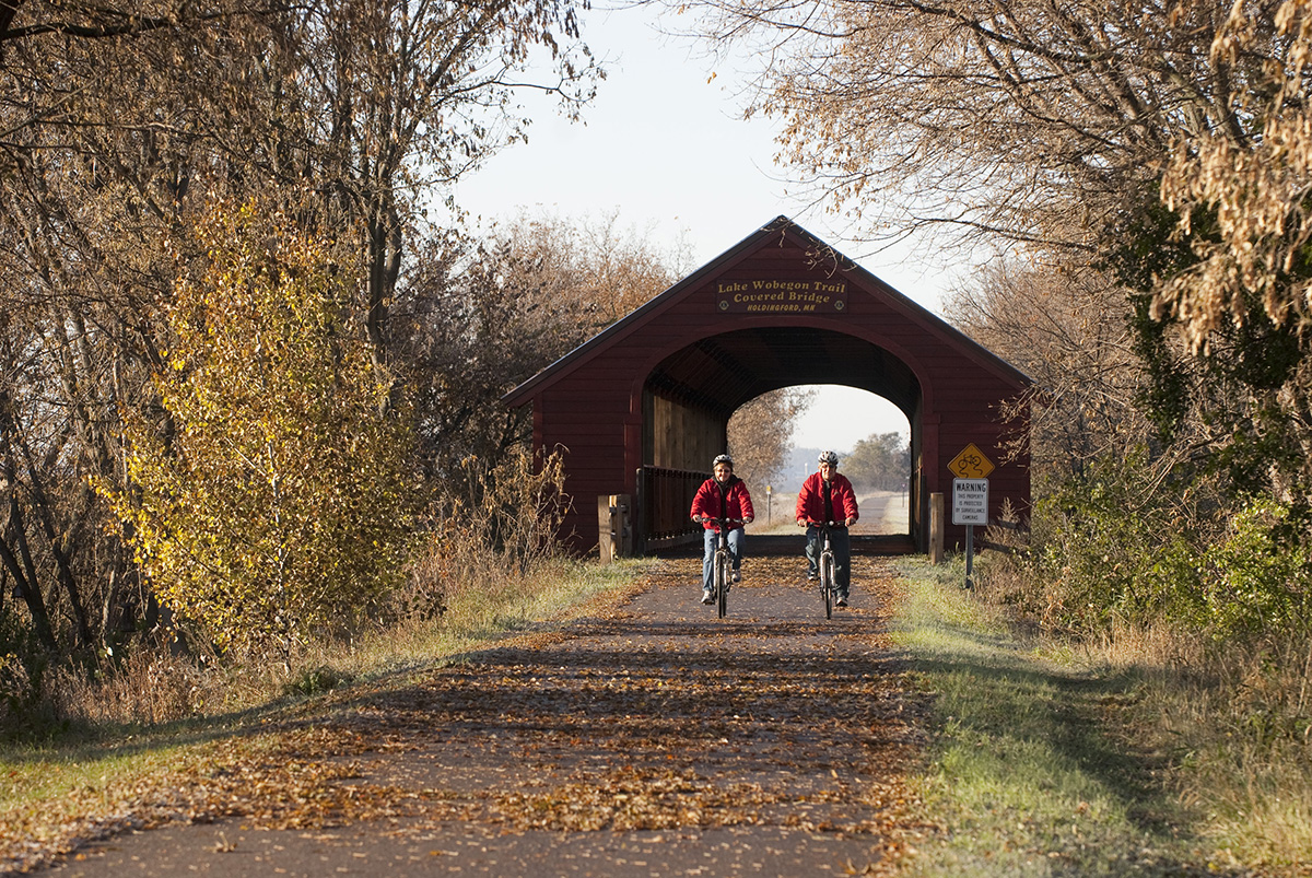 2 bicyclists ride on a paved trail with covered structure/tunnel behind them during fall season