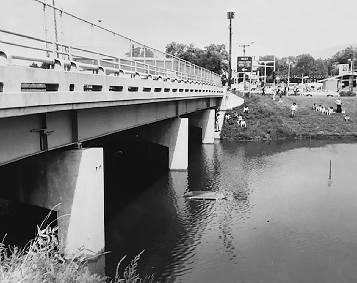 1987 black and white photo of concrete bridge, rising water and people sitting on river bank