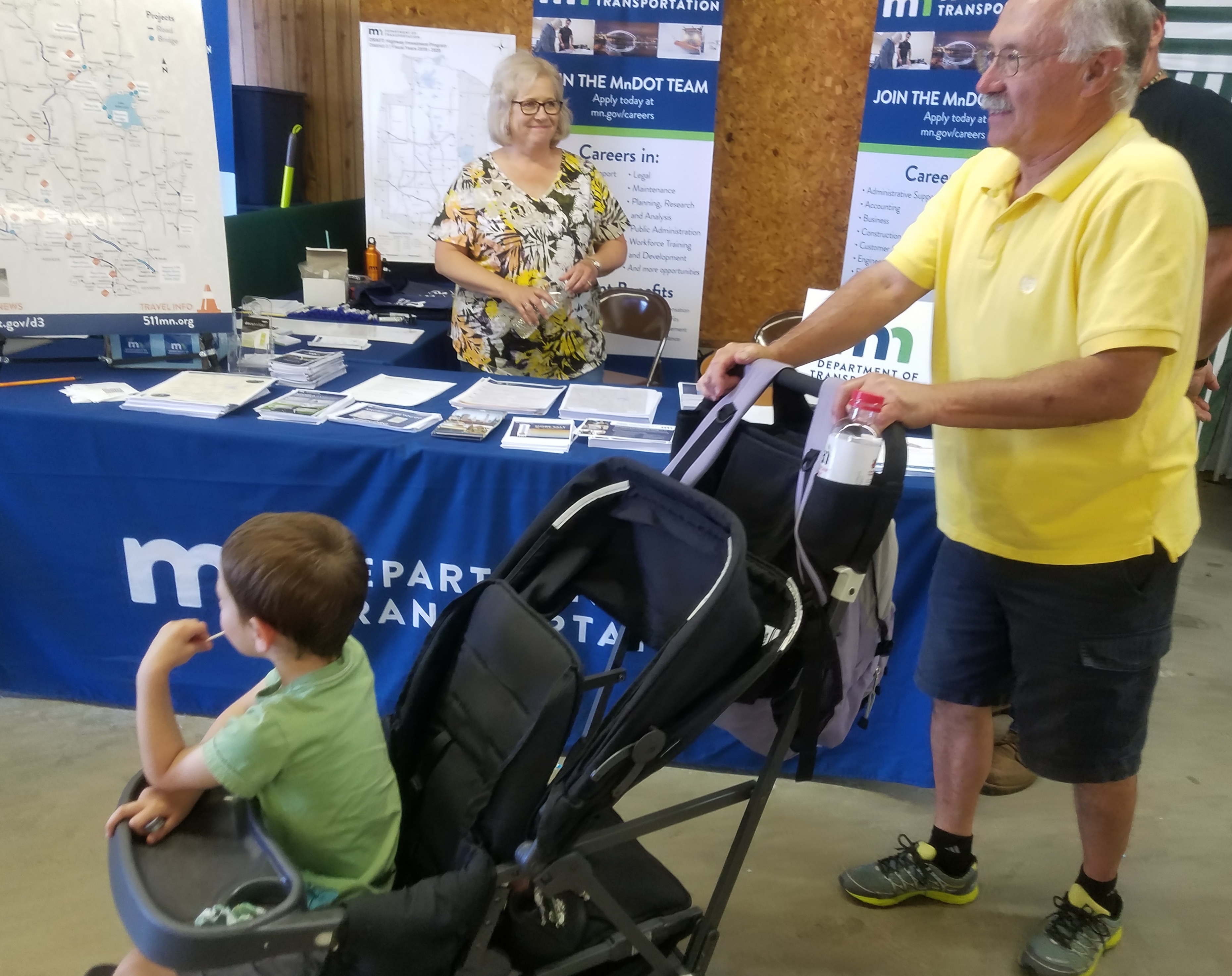 Cindy Aselson looks on as a man pushes a stroller past the MnDOT booth at the crow wing county fair