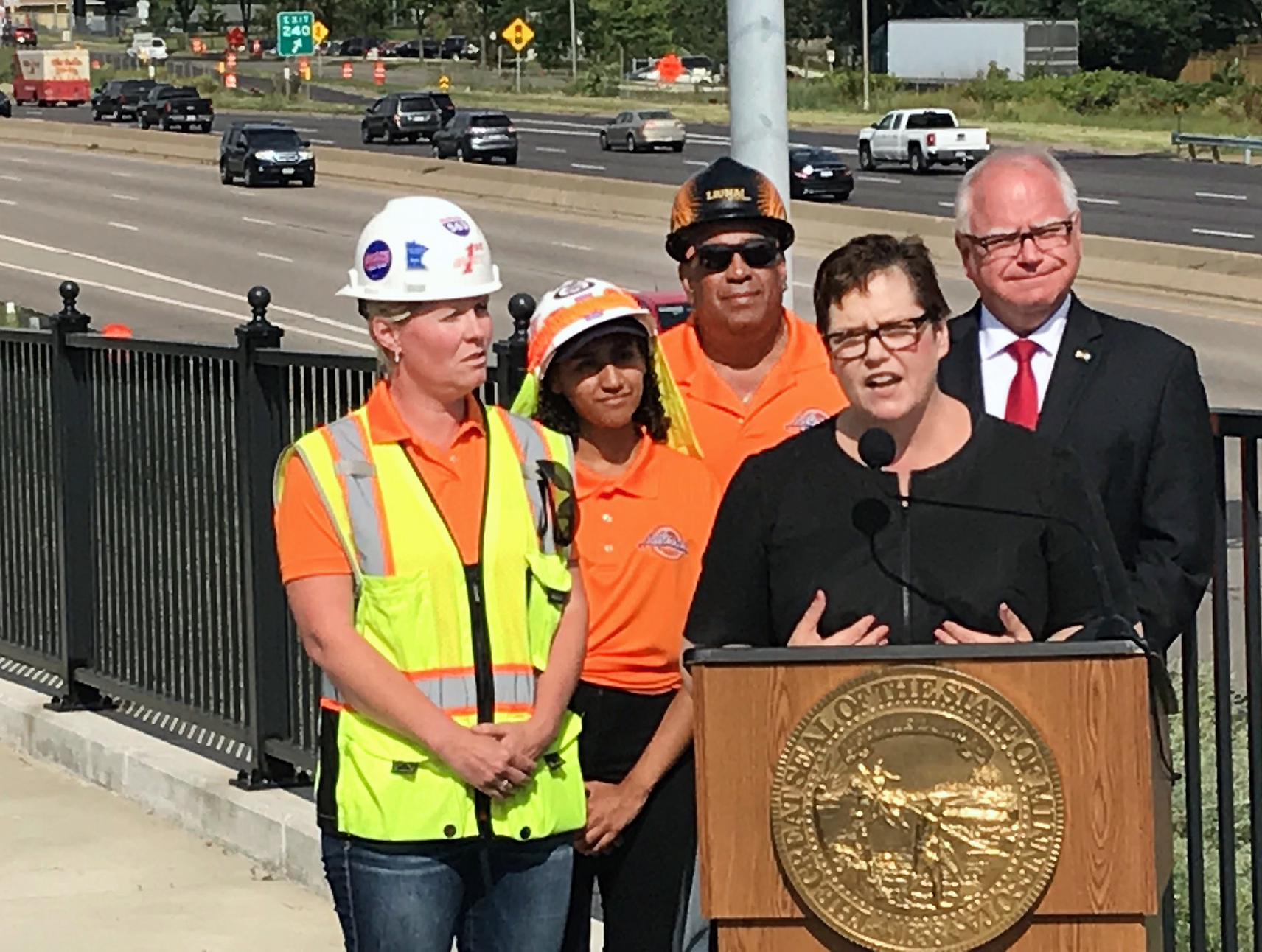 Commissioner maragaret anne kelliher speaking at a podium. Gov. tim walz is to her left, three other people are two her right, and a busy section of freeway is behind her.