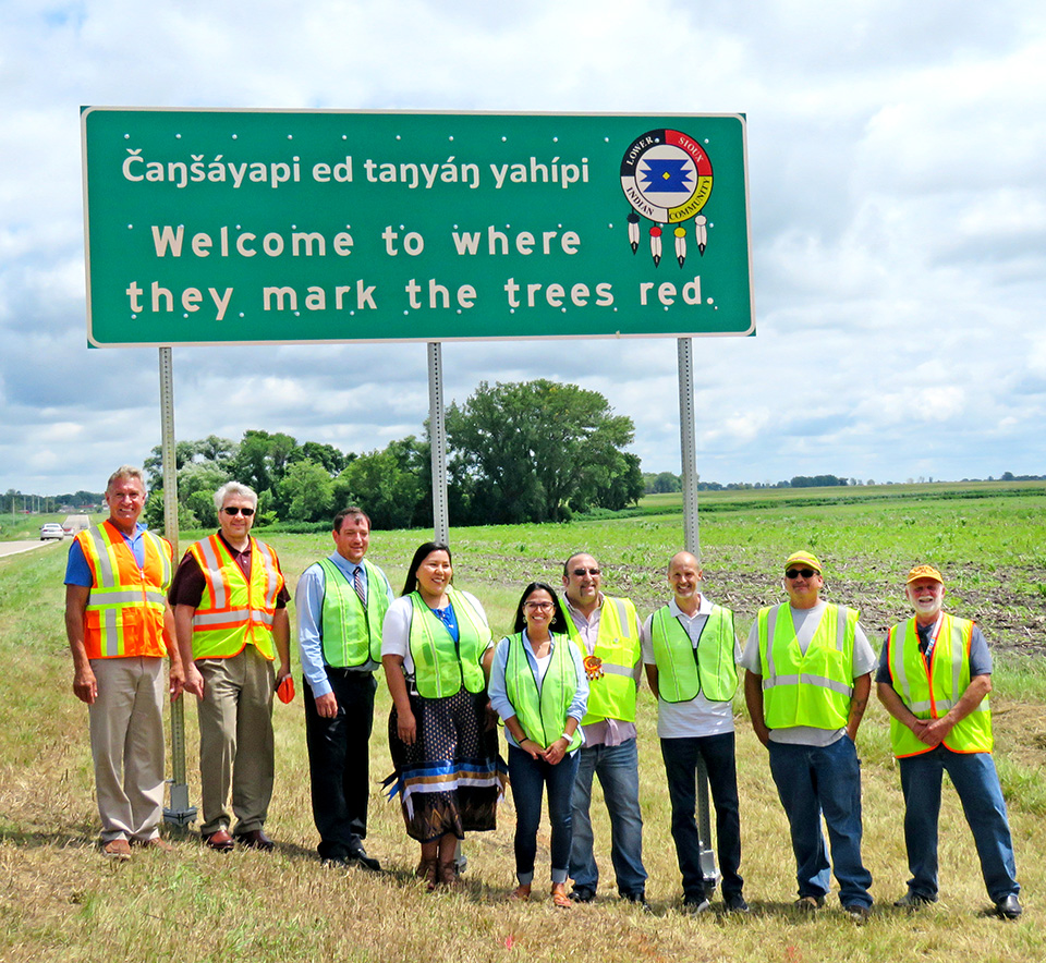 A groiup of people in safety vests are standing in front of a sign with Dakota and English words on it. The English words on the sign say welcome to where they paint the trees red.