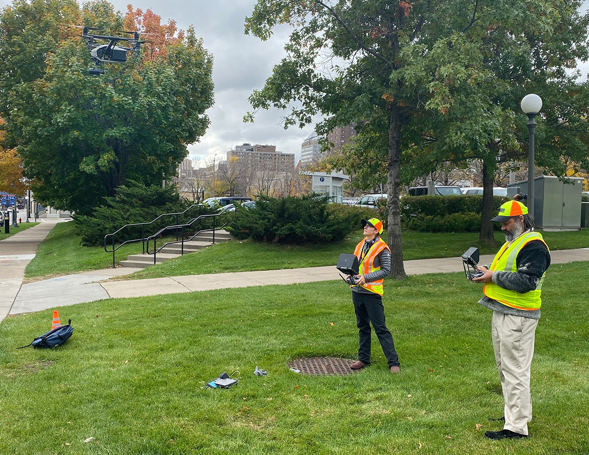Photo: two people in yellow safety vests look up at a low-flying drone they are operating
