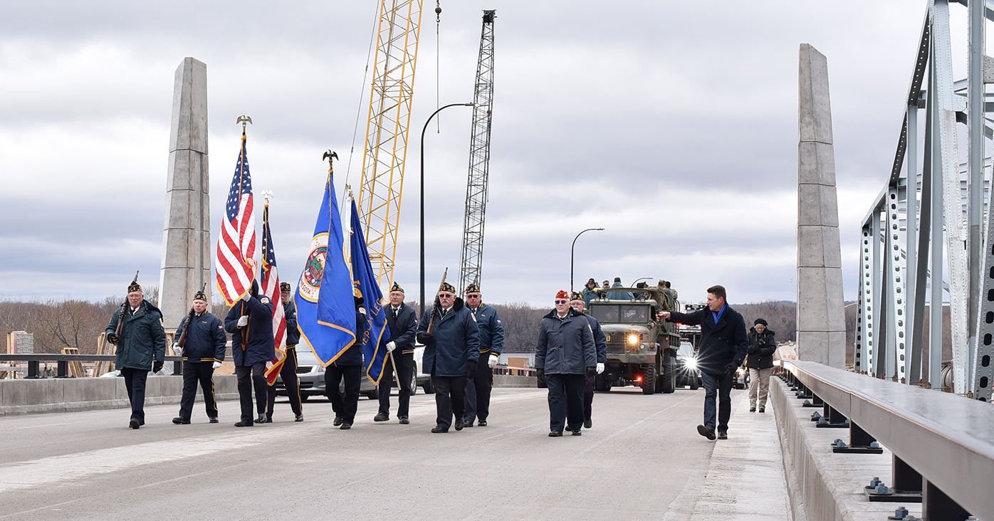 This picture shows a line of men carrying state and U.S. flags in a line while walking across the newly completed bridge. Several vintage military vehicles are behind the men, also crossing
