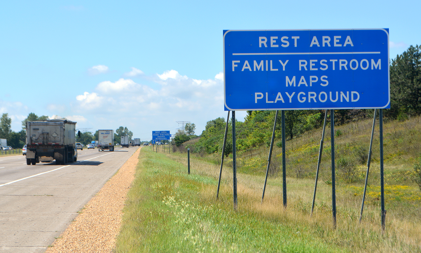 This photo shows a highway sign which has the words Rest Area on the top line, listing ameneties family restroom, maps and playground written underneath that.