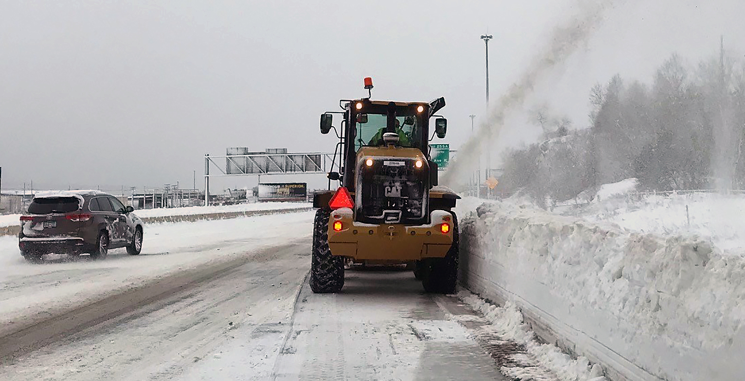 This photo shows a large snowblower blowing snow on the side of I-35. The snow to the right of the blower is close to four feet tall.