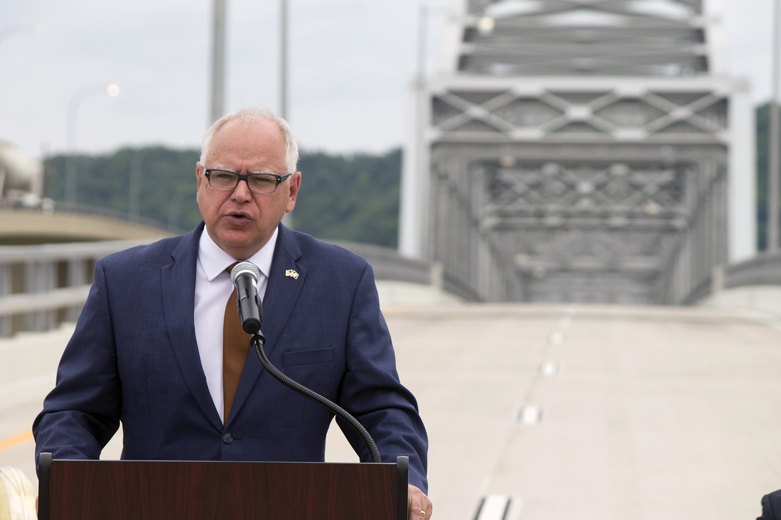 Minnesota governor Tim Walz speaking at a podium with the bridge in the background