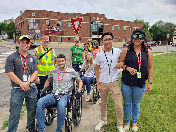 Interns and staff standing at a street corner and looking at the camera. Two interns are in wheelchairs, and one is weaing vision impairment goggles. These devices are meant to simulate what it is like for people with disabilities when they use the street to travel