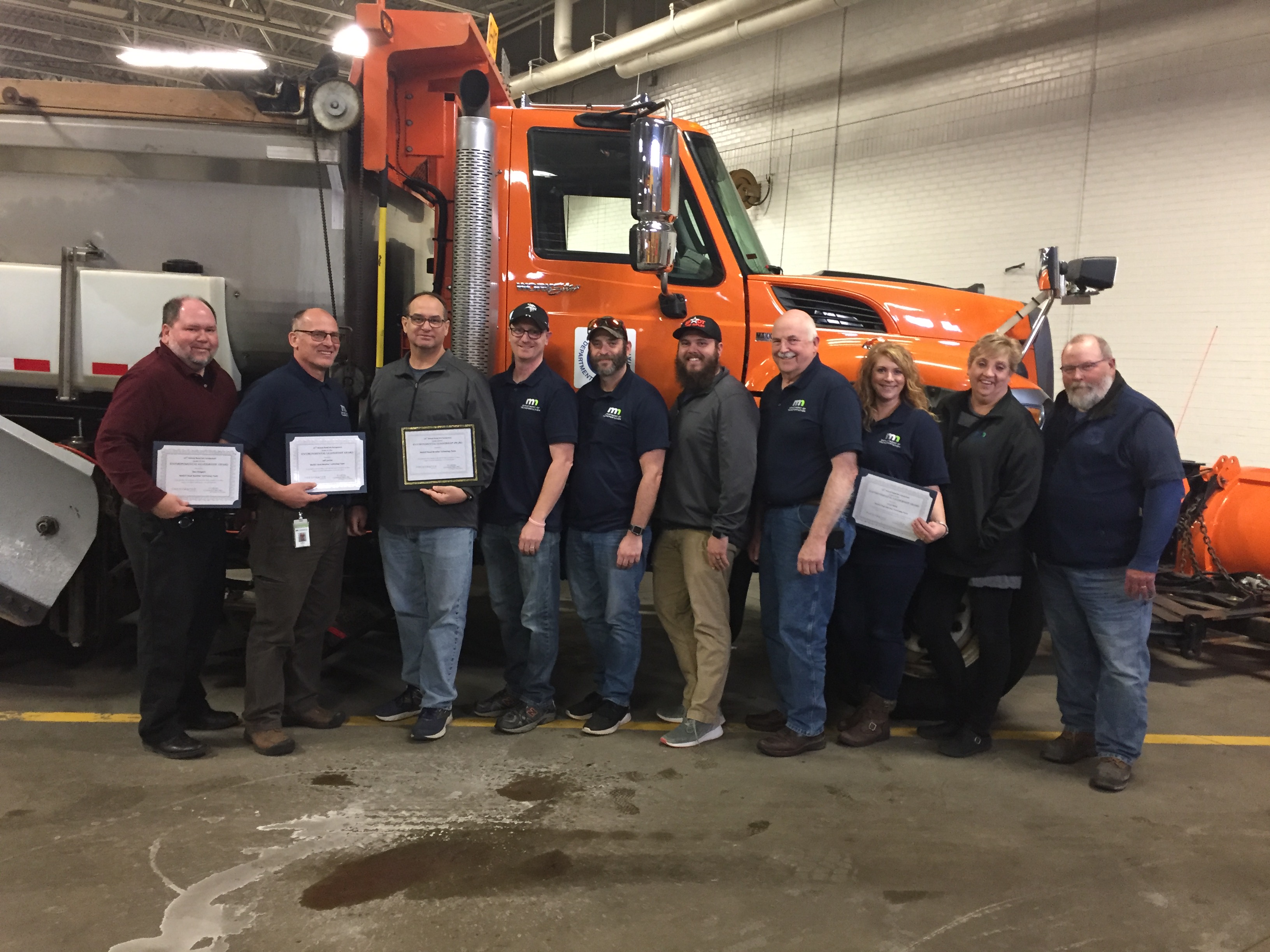 Nine people lined up in front of a plow truck, with four holding certificates