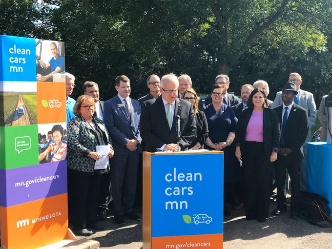 Gov. Walz speaking at a podium outside on a sunny day. A sign that says Clean Cars MN is mounted on the front of the podium