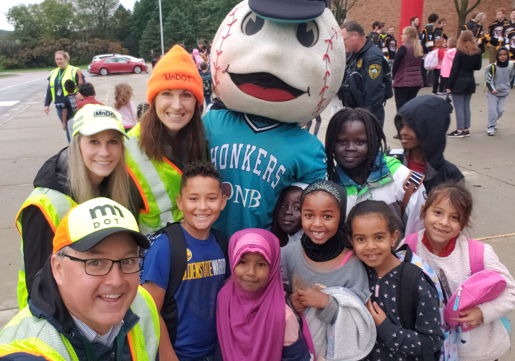 Three MnDOT employees and a group of school children pose on a sidewalk outside of the school. A mastoc with a large baseball for a head is also pictured with them.
