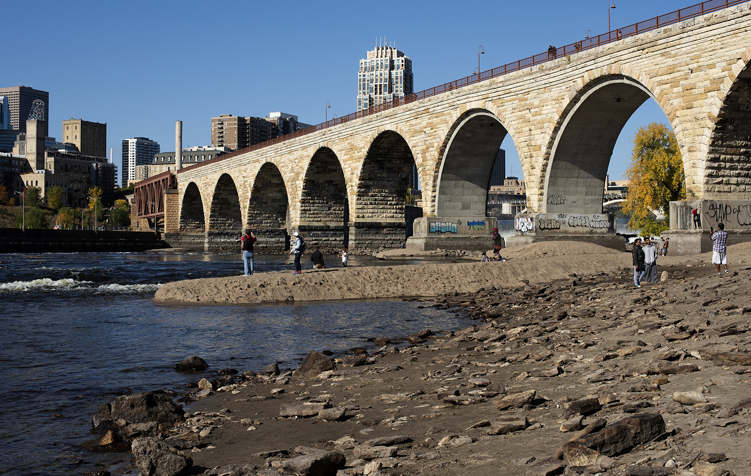 Photo: people walking around on the exposed mud left by lower water levels in the Mississippi River by the Stone Arch Bridge in Minneapolis.
