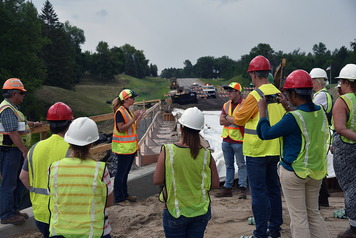 Photo: A group of people in safety vests listening to a MnDOT employee speaking near a work site